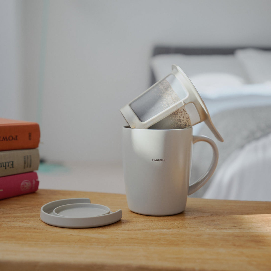 Pouring hot water from kettle into hario ceramic tea and coffee mug with infuser filter on wooden bedroom table