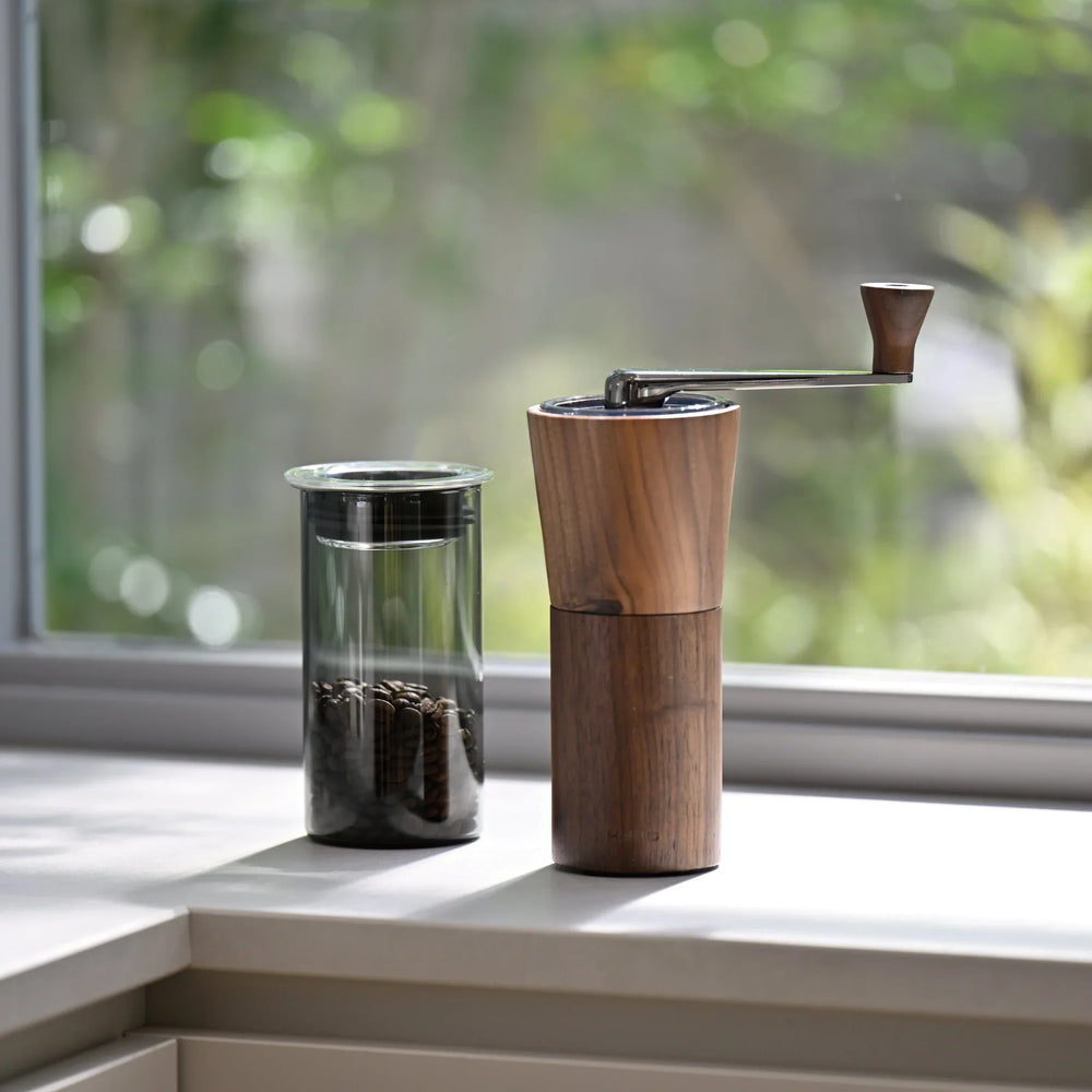 Placing hario walnut wood coffee grinder next to glass canister filled with coffee beans on sunny windowsill kitchen