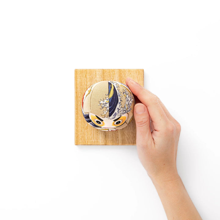 Fabric daruma ornament with bold Kabuki style face and wave pattern set on a small wooden display stand