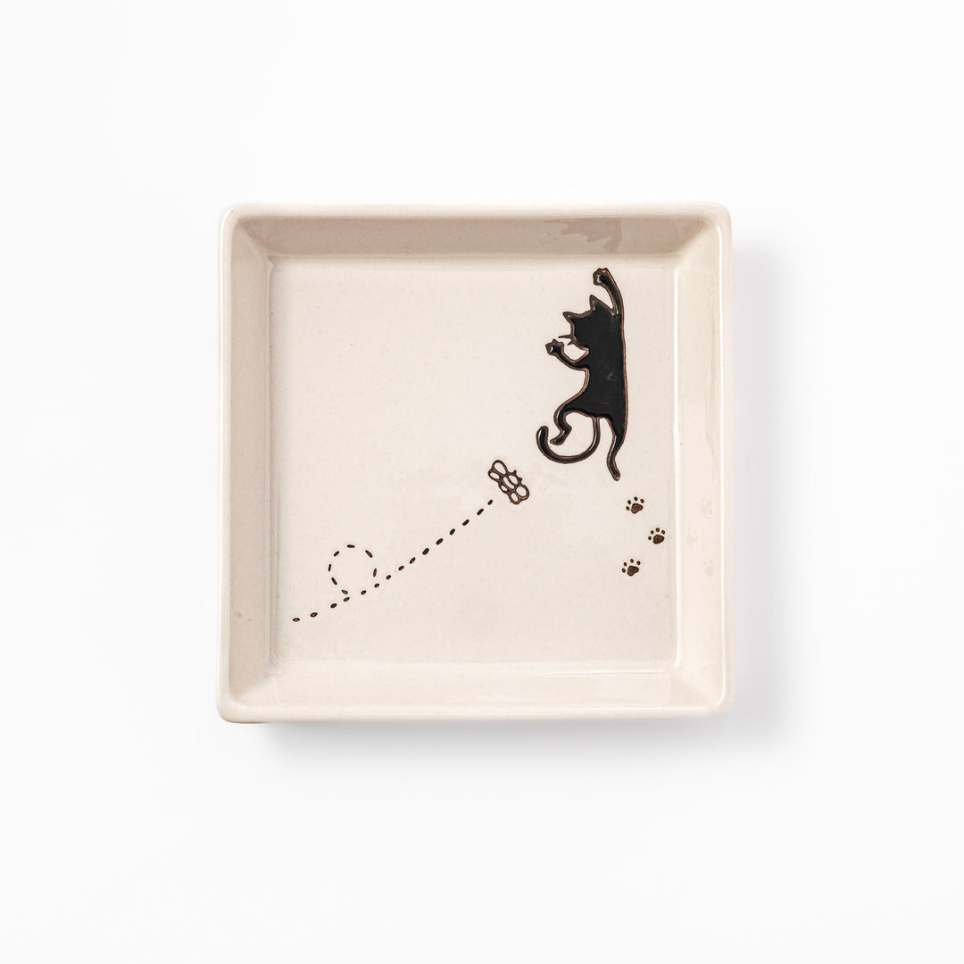 Square beige plate featuring a playful black cat chasing a butterfly, with paw prints and a dotted flight path.