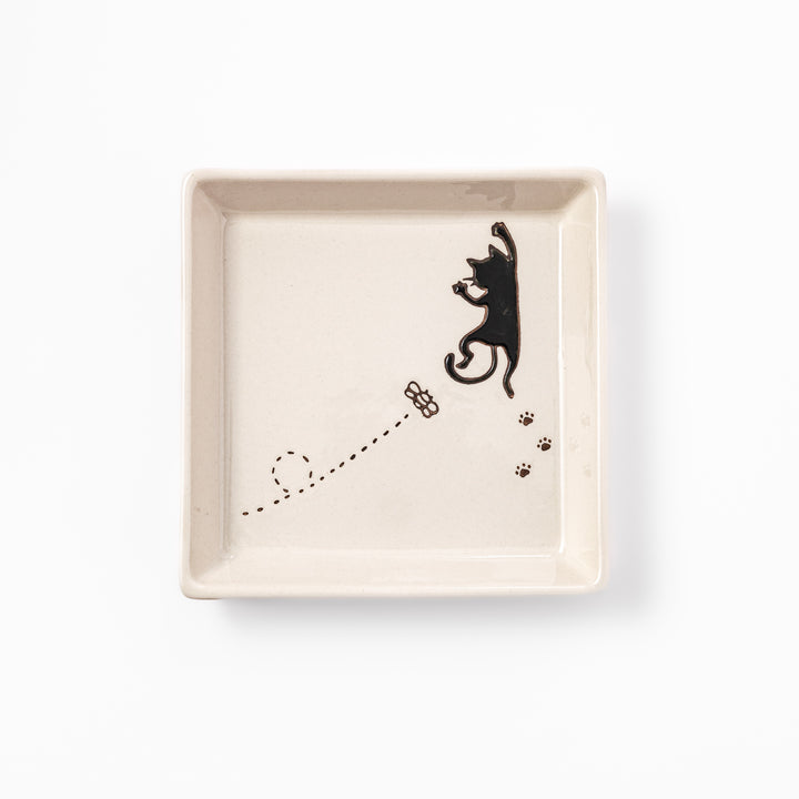 Square beige plate featuring a playful black cat chasing a butterfly, with paw prints and a dotted flight path.