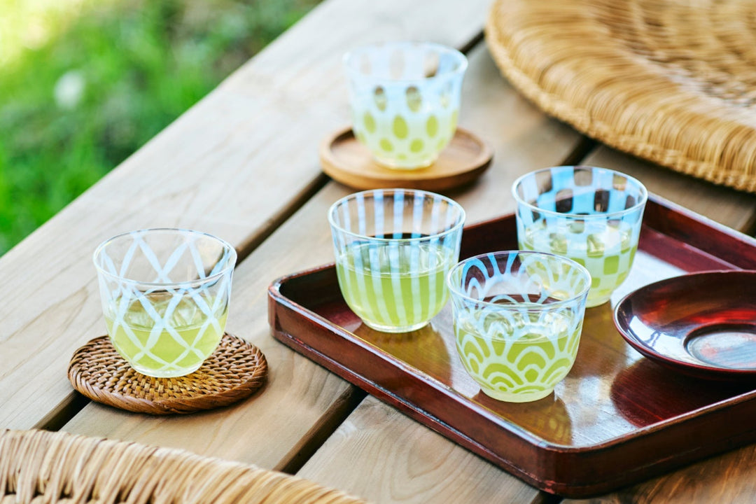 Patterned Japanese glass cups filled with green tea on a wooden outdoor table with a tray and woven coasters