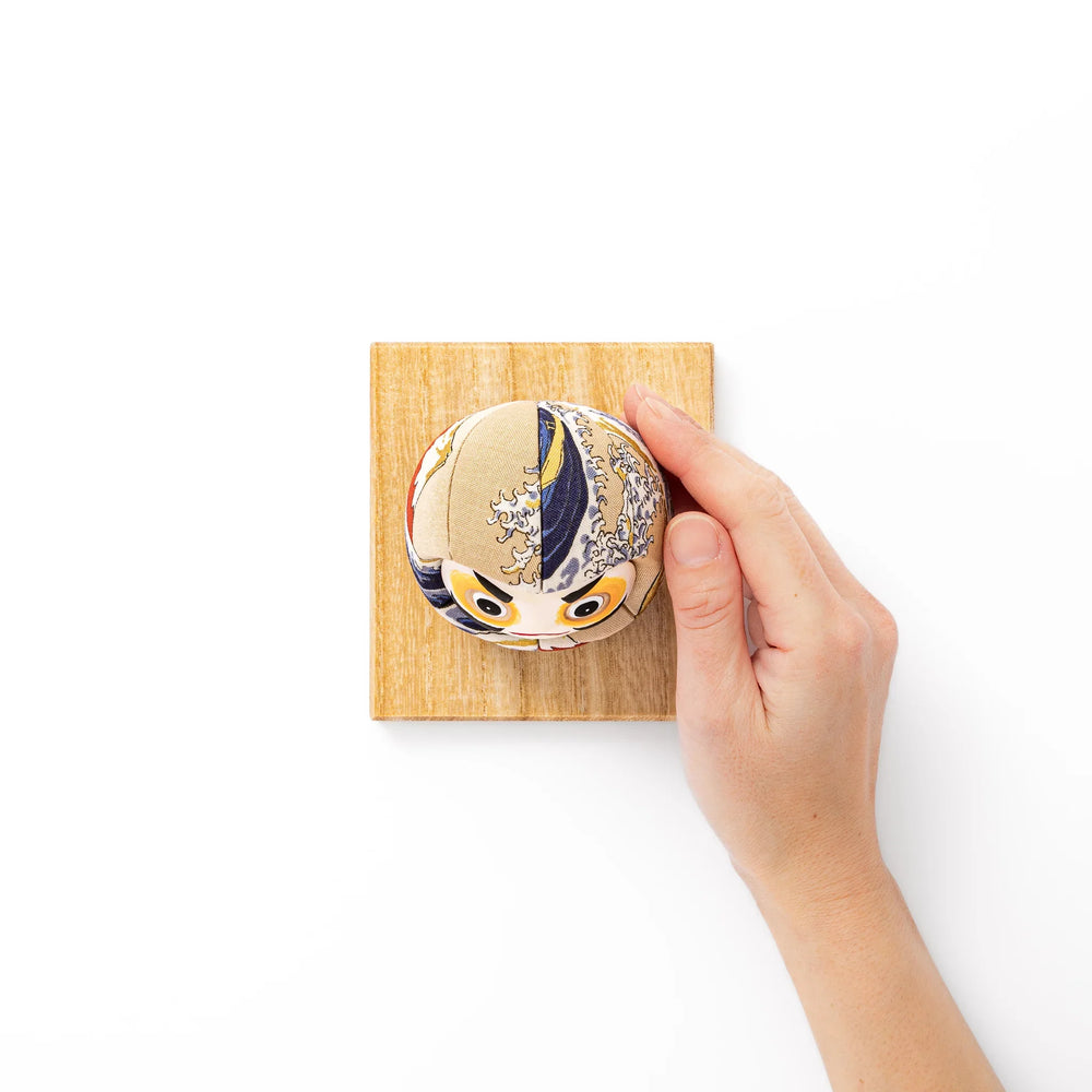 Fabric daruma ornament with bold Kabuki style face and wave pattern set on a small wooden display stand