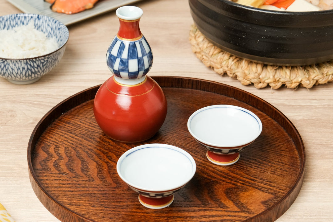 Japanese sake set with red and blue porcelain tokkuri and cups on a round wooden tray with a meal setting in the background
