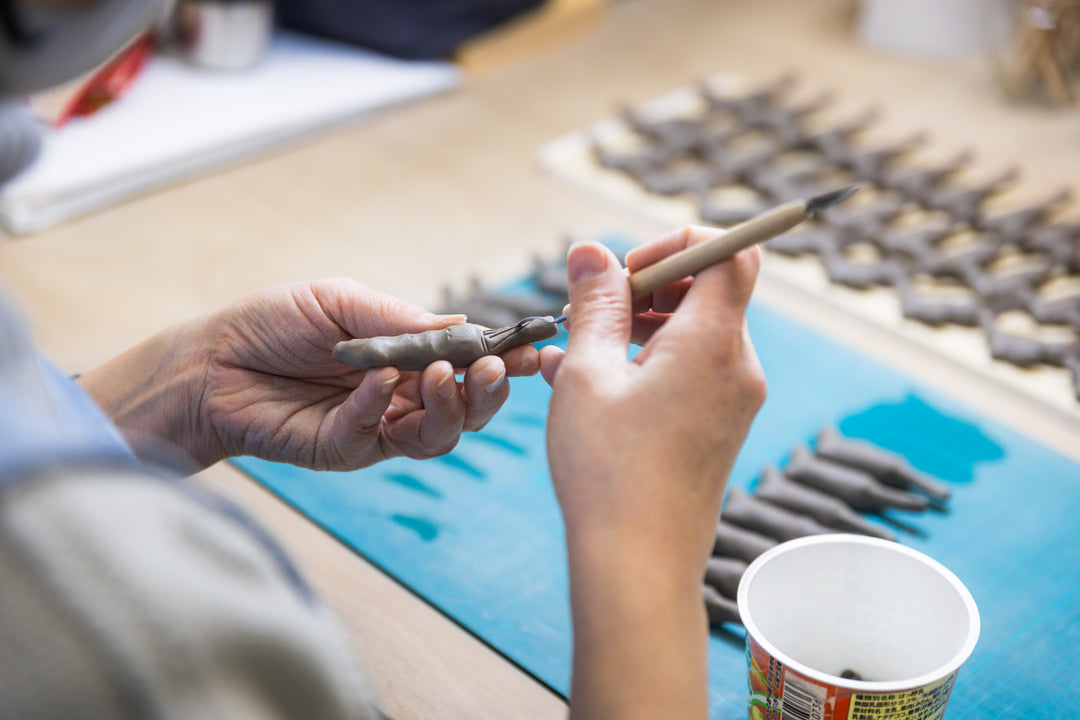 Clay artisan shaping a small figurine by hand, using a carving tool with rows of unfinished pieces drying on the table.