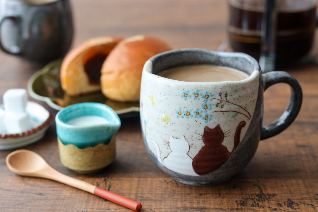 Cozy ceramic mug with hand painted cats and flowers filled with coffee on a wooden table with pastries and milk nearby