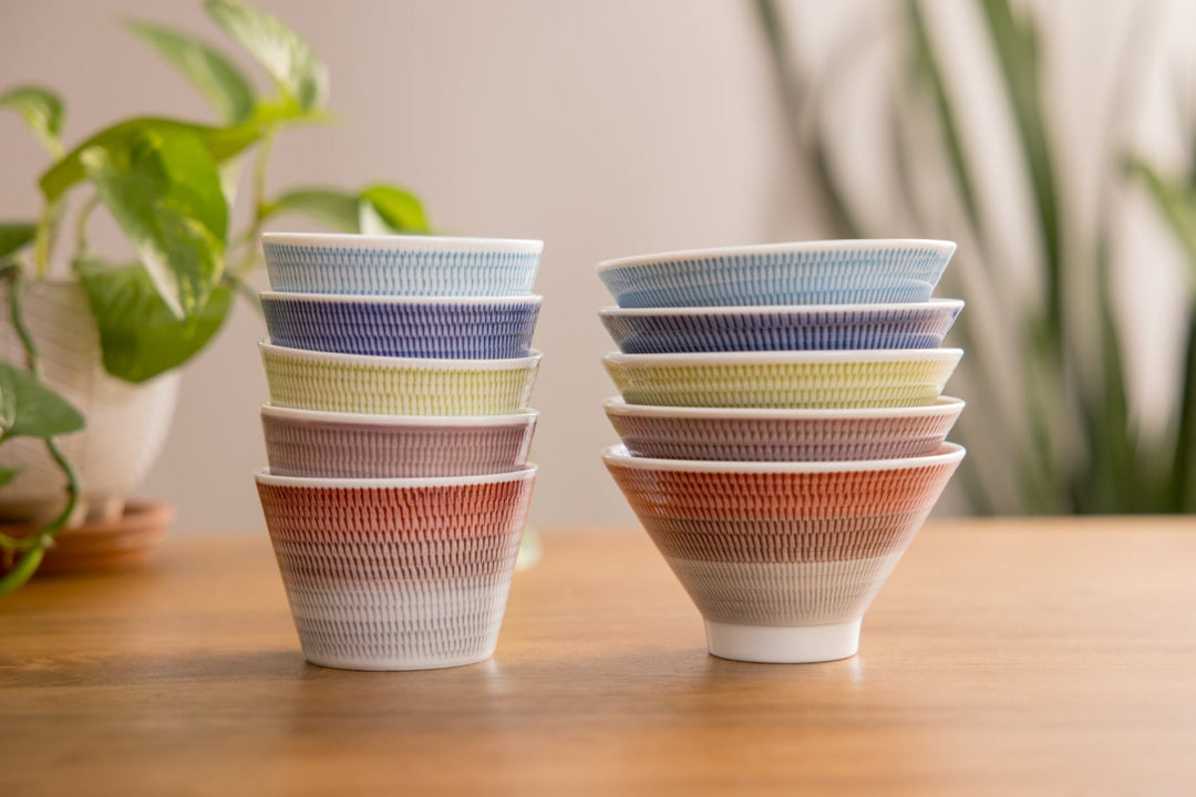 Colorful stack of small Japanese bowls with fine vertical patterns displayed on a wooden table with green plants in the background.