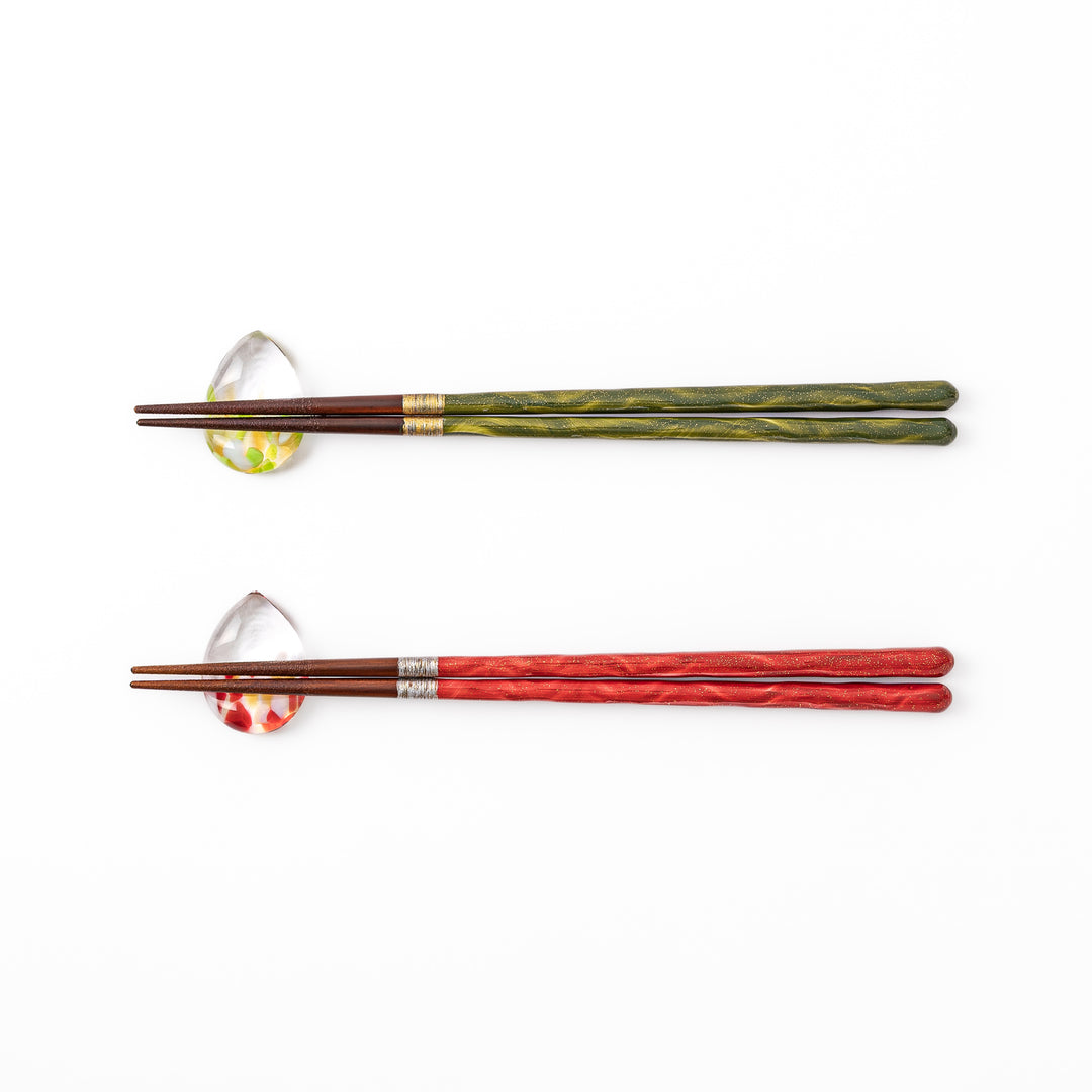 A pair of green and red Japanese lacquer chopsticks next to two teardrop shaped colorful translucent glass chopstick rests