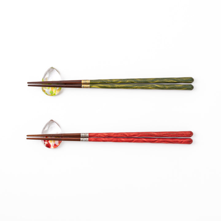 A pair of green and red Japanese lacquer chopsticks next to two teardrop shaped colorful translucent glass chopstick rests