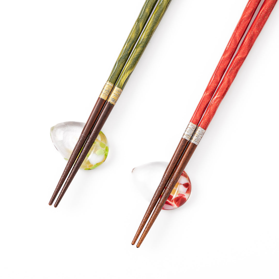 A pair of green and red Japanese lacquer chopsticks next to two teardrop shaped colorful translucent glass chopstick rests
