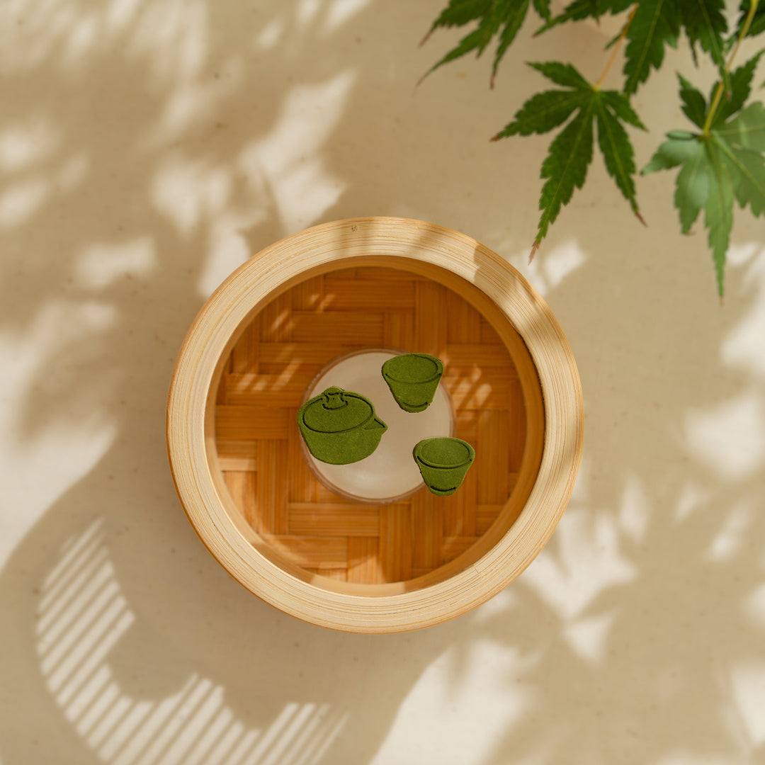 Green Japanese matcha pressed incense shaped like a teapot and teacups inside a round bamboo incense warmer