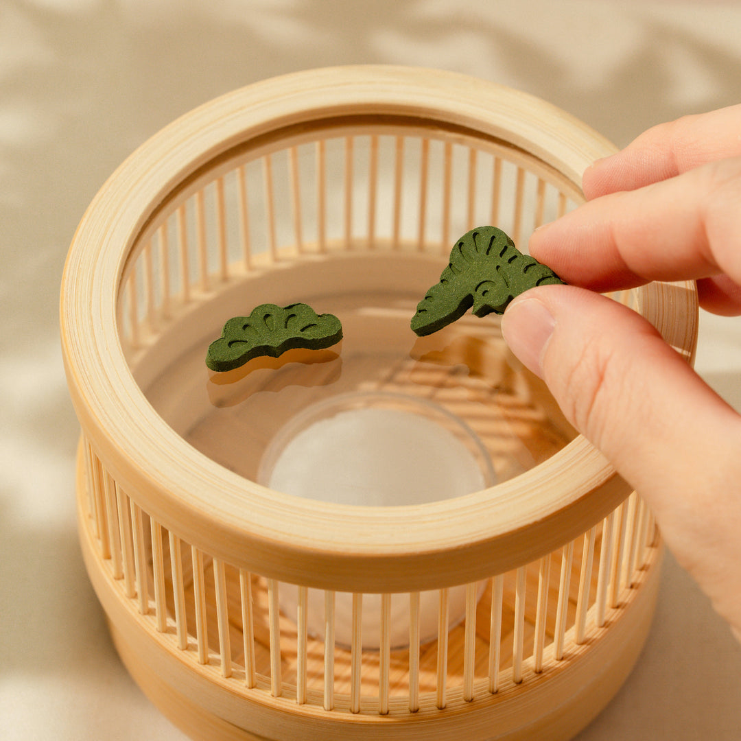 A hand placing green pine shaped matcha pressed incense into a round bamboo incense burner