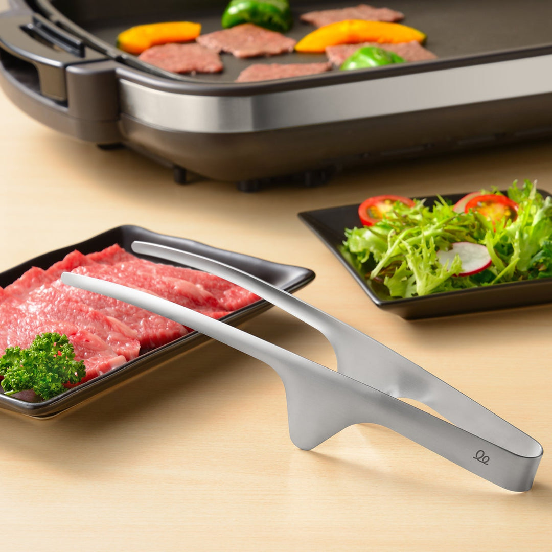 Japanese stainless steel tabletop BBQ tongs resting on a table with elevated tips to keep the surface clean and hygienic