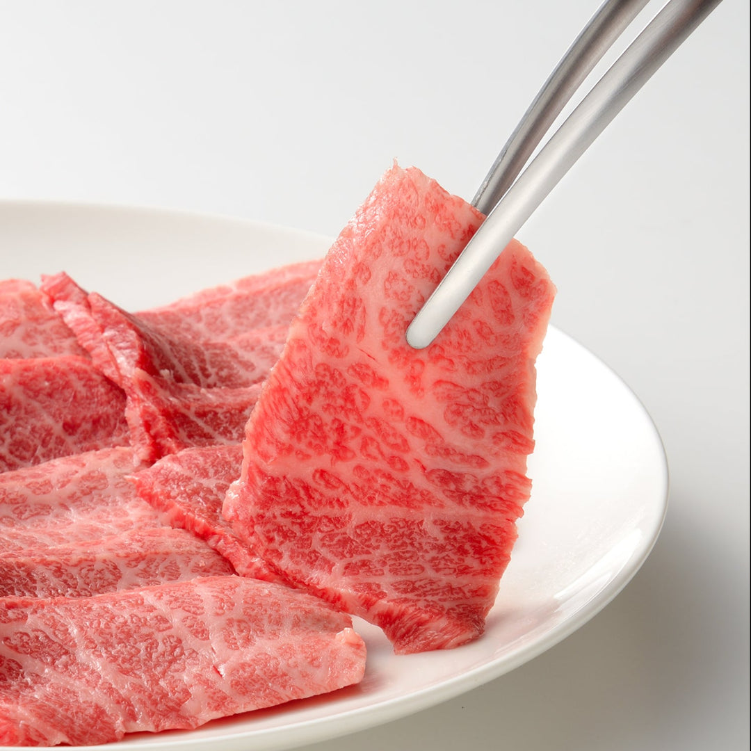 Japanese stainless steel BBQ tongs picking up a slice of raw marbled wagyu beef from a white plate for grilling
