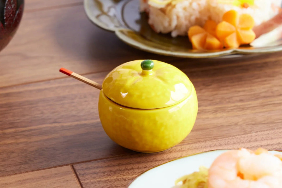 Bright yellow ceramic yuzu shaped condiment jar with lid and small spoon on a wooden dining table