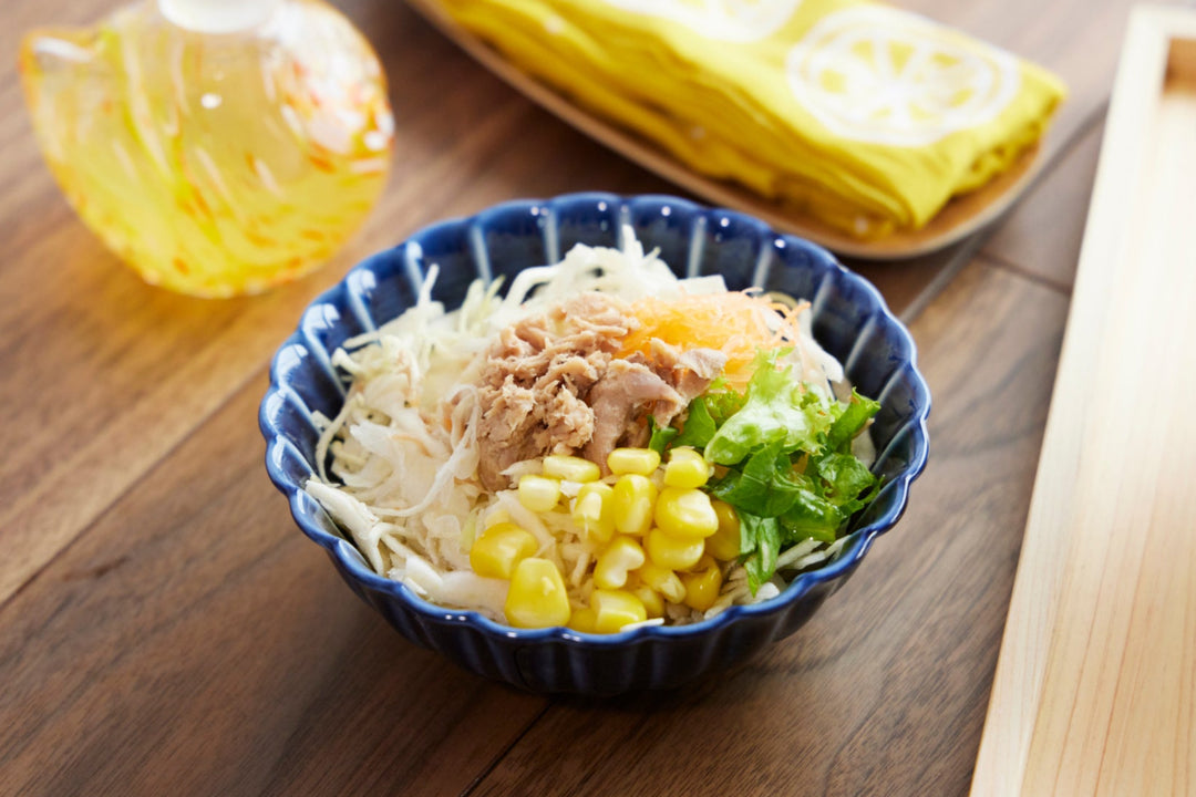 Deep blue scalloped Japanese bowl filled with shredded cabbage tuna corn and greens on a wooden table with a glass dressing bottle nearby