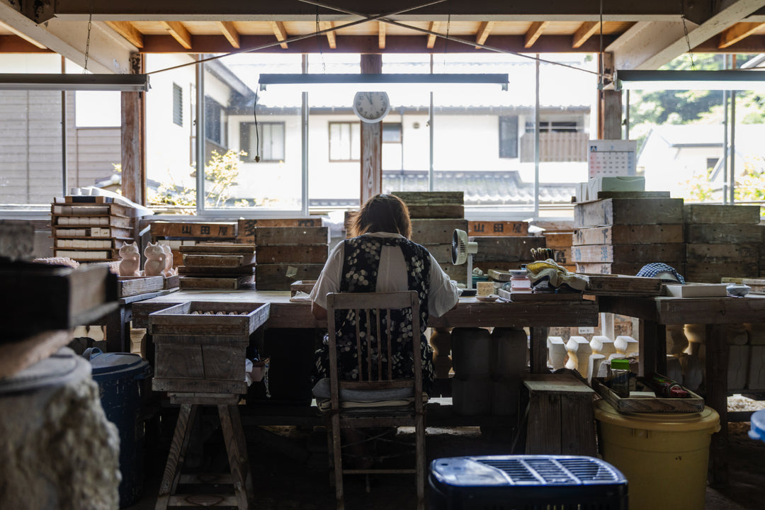 Japanese ceramic artisan working at a wooden table among molds and trays in a bright, traditional workshop.