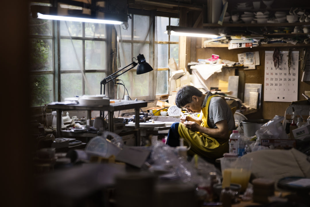 A craftsman in a yellow apron carefully carving a ceramic piece at a wooden worktable inside a traditional pottery studio.