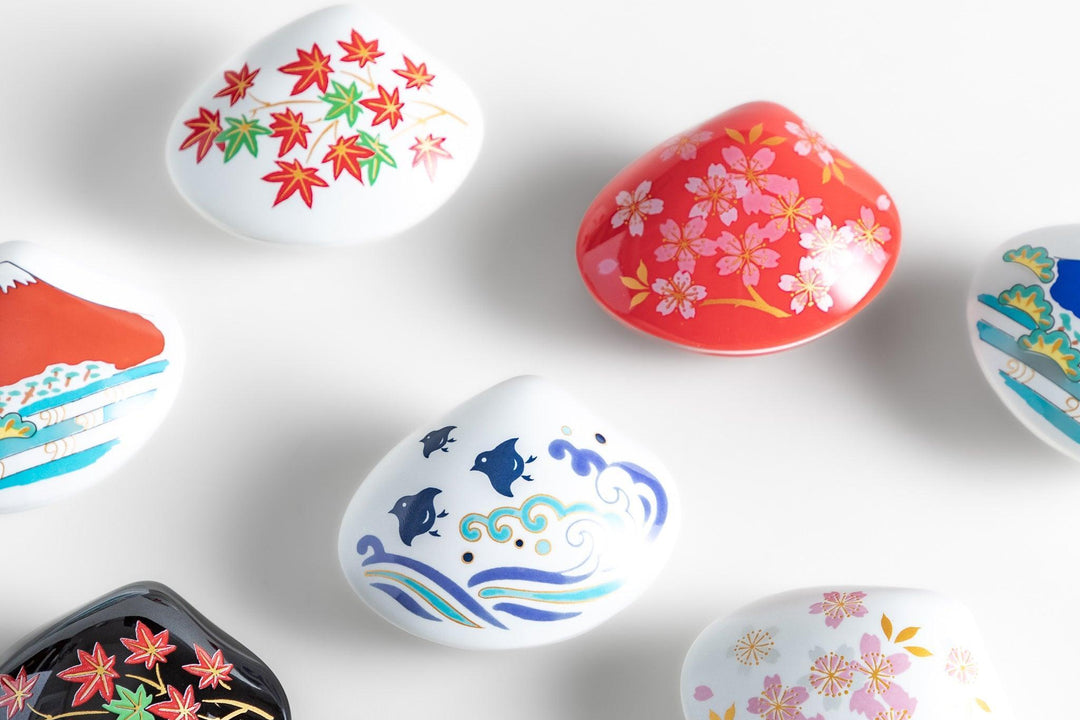 Colorful Japanese ceramic clam shape dishes with seasonal motifs including sakura maple waves and Mount Fuji on white background.