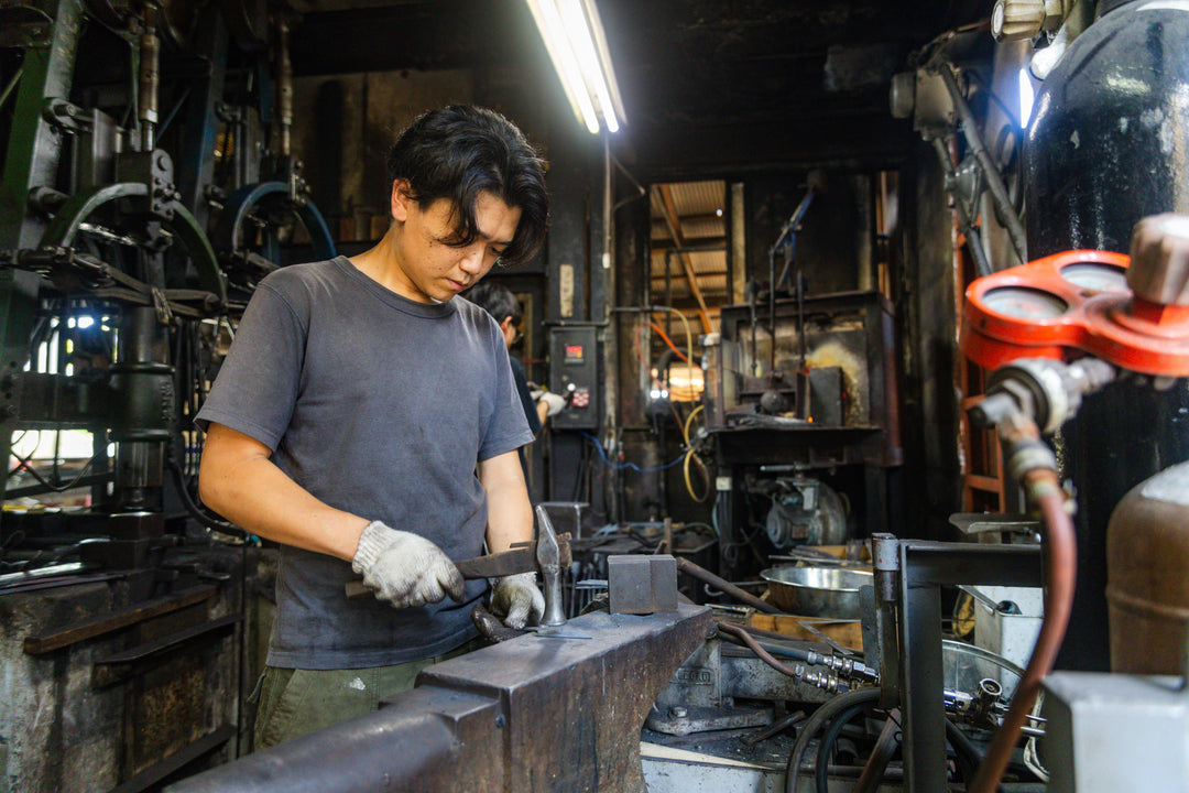 A metalworker in a black shirt hammering heated steel on an anvil inside a busy traditional forging workshop.