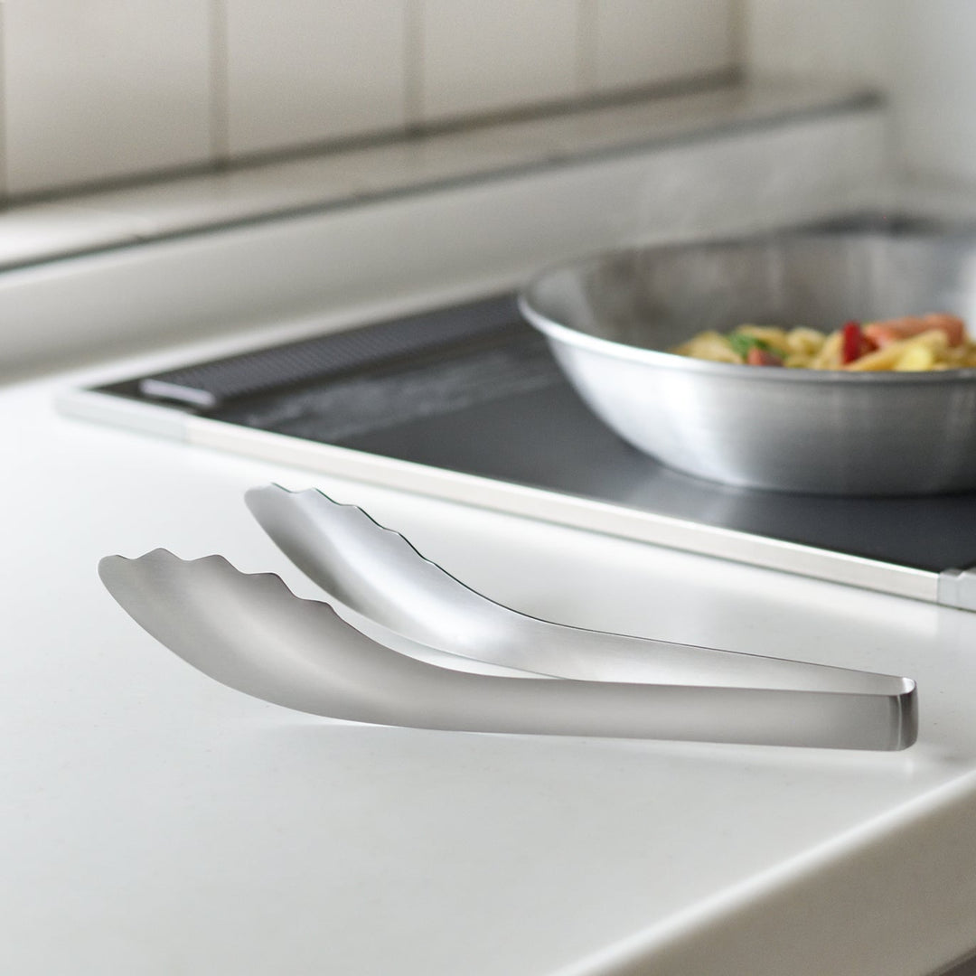 Japanese stainless steel noodle tongs resting on a white kitchen counter with elevated tips next to a steaming stir fry pan