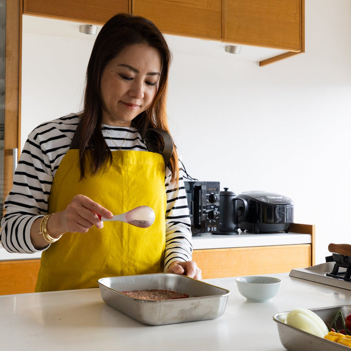 Nami using white spice maraca sifter to season raw steak meat inside metal tray on kitchen counter with yellow apron