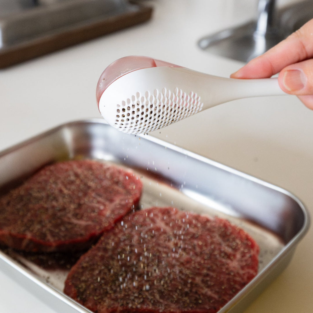 Nami shaking white spice maraca sifter to evenly distribute seasoning powder over raw beef steaks in metal tray tool