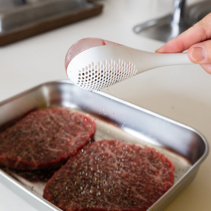 Nami shaking white spice maraca sifter to evenly distribute seasoning powder over raw beef steaks in metal tray tool