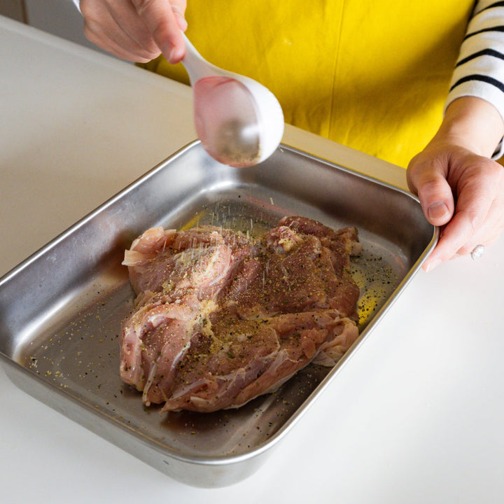 Nami sifting herb seasoning onto raw chicken meat in metal tray with white spice maraca sifter dispenser gadget