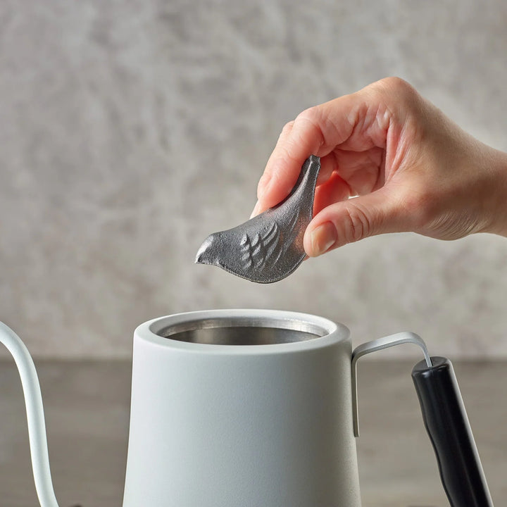 Person placing a bird shaped cast iron supplement into a white water kettle to enrich boiling water with iron