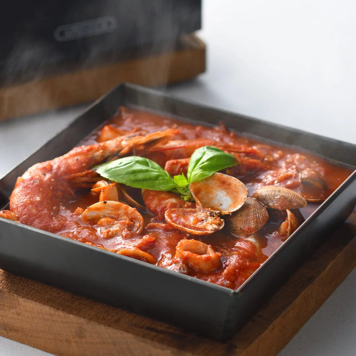 Steaming seafood tomato soup served in a square compact deep frying pan resting on a wooden trivet