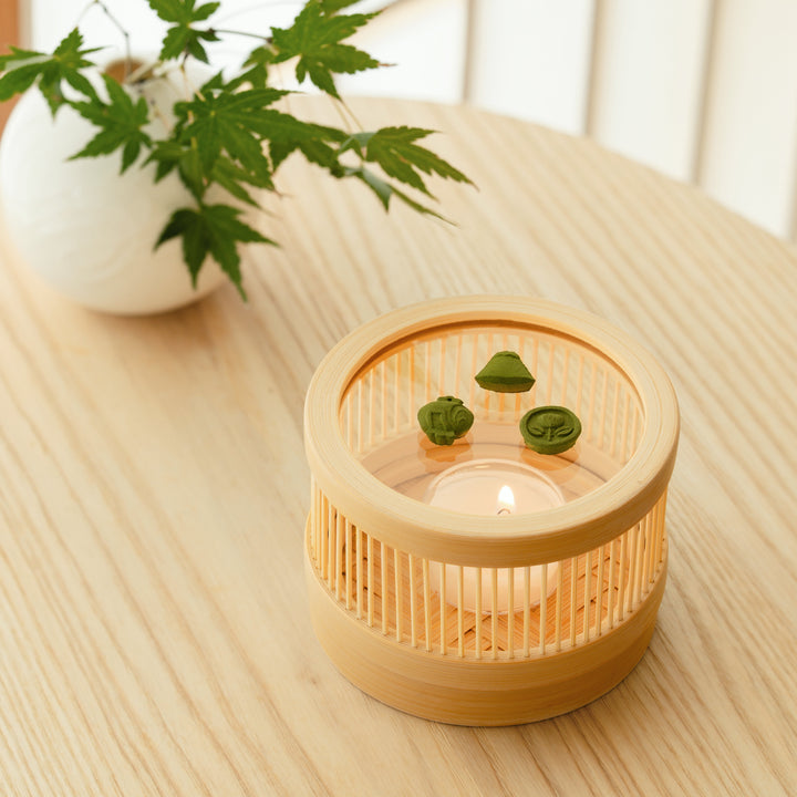 A round bamboo incense warmer with green matcha pressed incense on a wooden table with green maple leaves