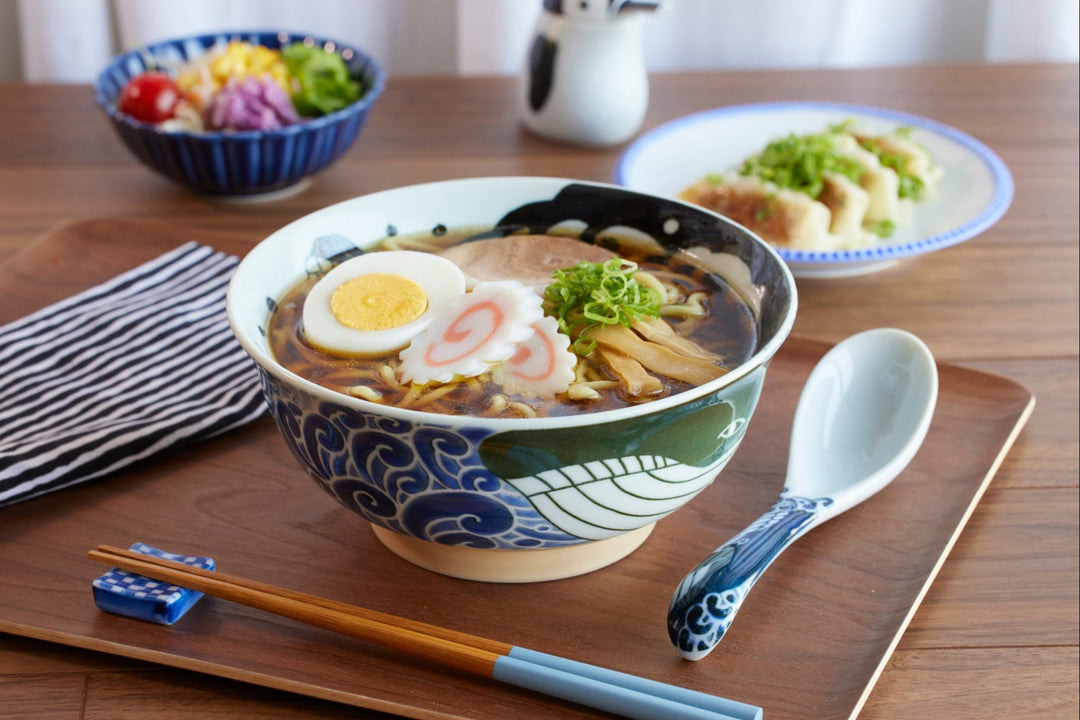 Japanese ramen bowl with wave motif filled with ramen, served with chopsticks, spoon, salad, and gyoza.