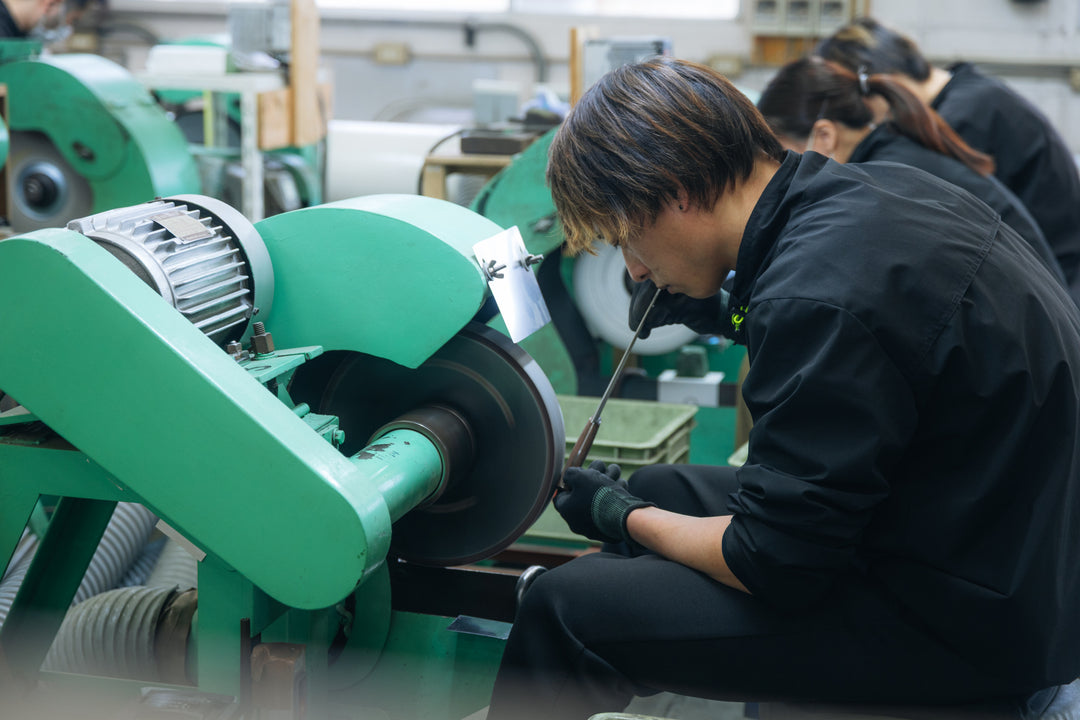 A craftsman in black workwear polishing a knife blade using a large green grinding machine inside the factory.