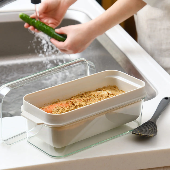 Japanese nukadoko fermentation box with rice bran bed by the sink, shown with spatula and cucumber being washed for pickling.