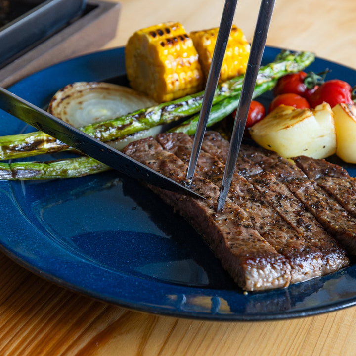 Nami using japanese carving knife and fork to slice grilled steak on blue dinner plate with barbecue vegetables