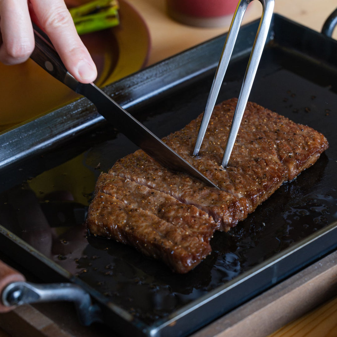 Japanese carving knife and fork slicing grilled steak meat on rectangular iron griddle plate kitchenware bbq