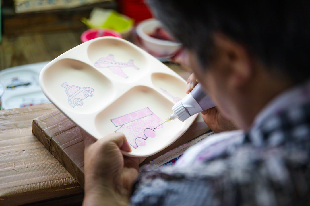 Artisan hand-coloring a children’s divided plate with playful designs, adding details using a fine applicator tool.