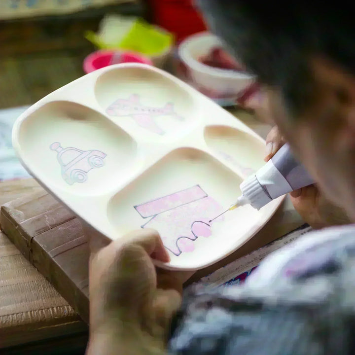 Artisan hand-painting a children’s divided plate with pink vehicle designs, showing careful brushwork and craftsmanship.