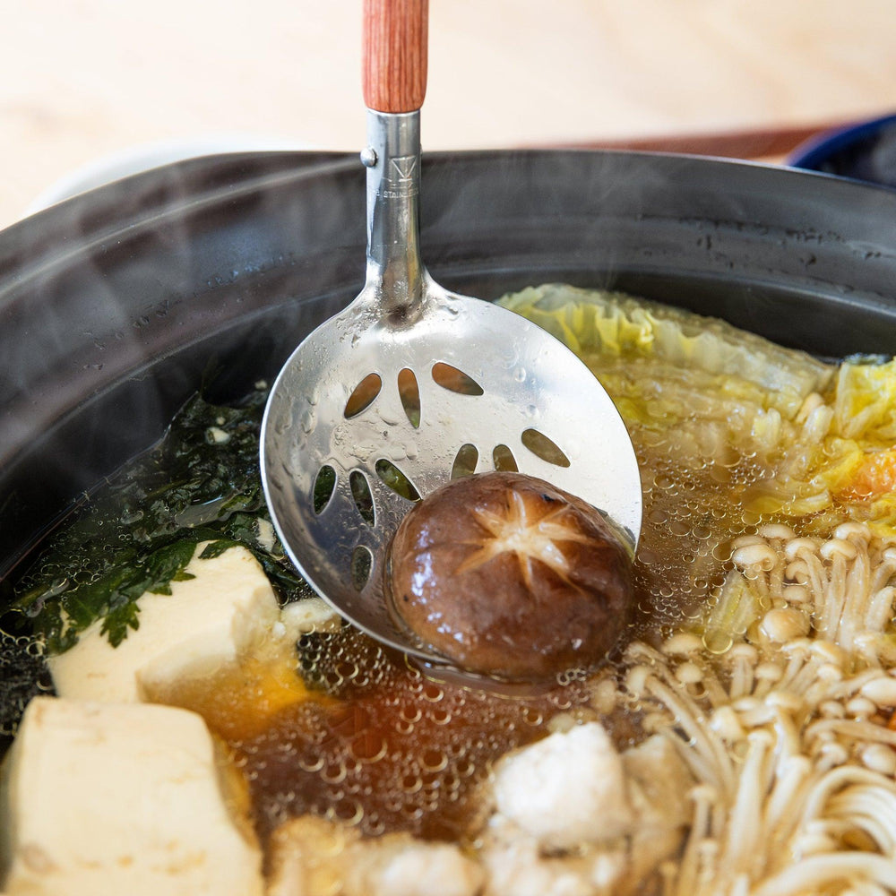 Perforated stainless steel soup ladle with wooden handle lifting shiitake mushroom from donabe hot pot