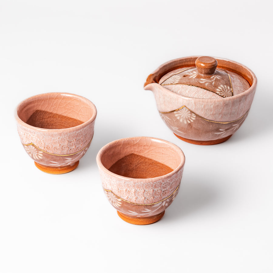 Three piece ceramic tea set with a soft pink crackled glaze and floral design, featuring a spouted teapot and two matching cups with gold accents.