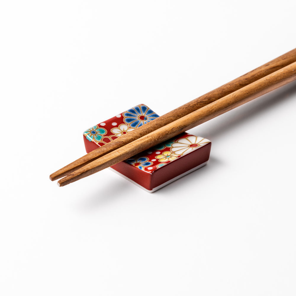 Rectangular Japanese ceramic chopstick rest with a red background decorated with colorful plum and chrysanthemum blossoms, perfect for elegant dining settings.