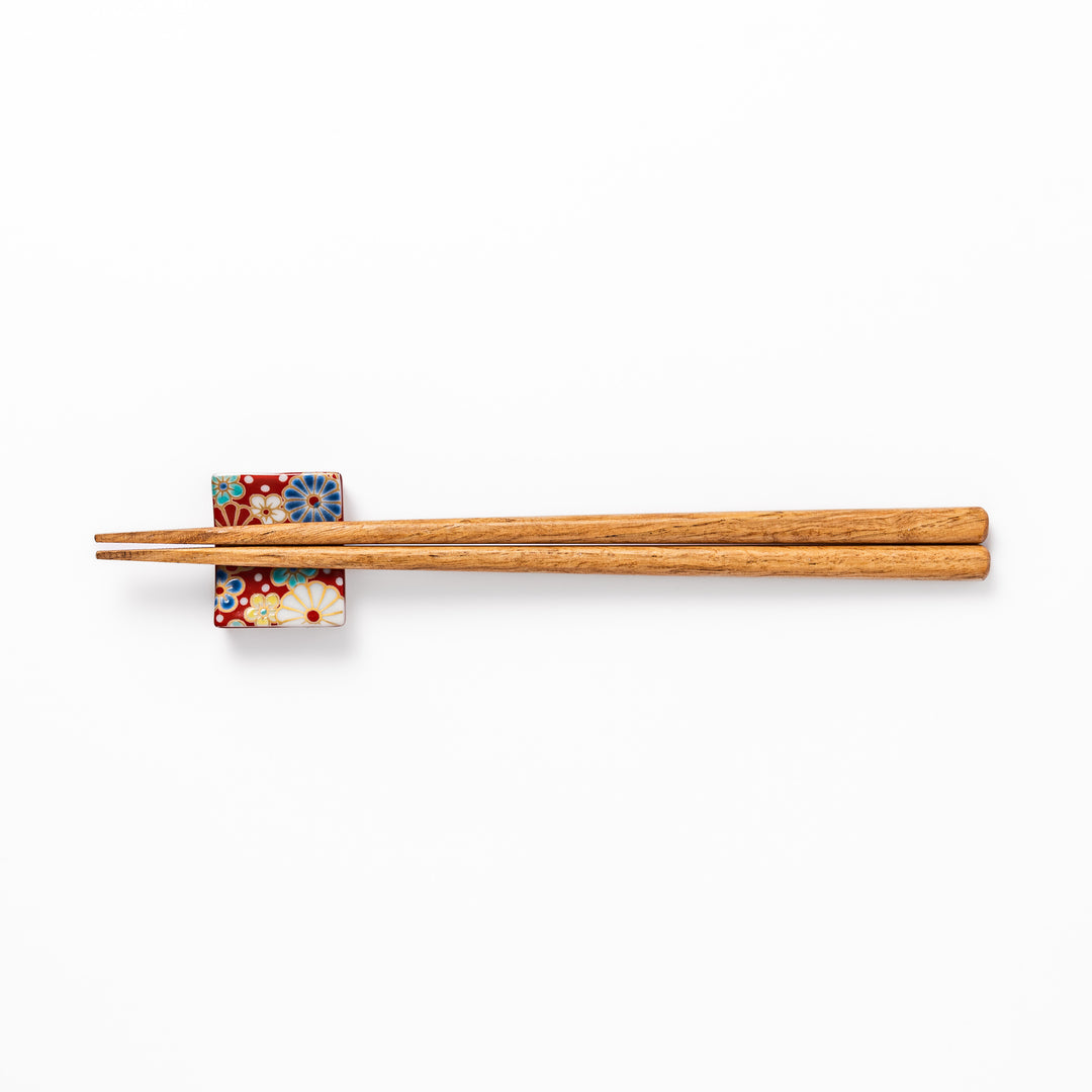Rectangular Japanese ceramic chopstick rest with a red background decorated with colorful plum and chrysanthemum blossoms, perfect for elegant dining settings.