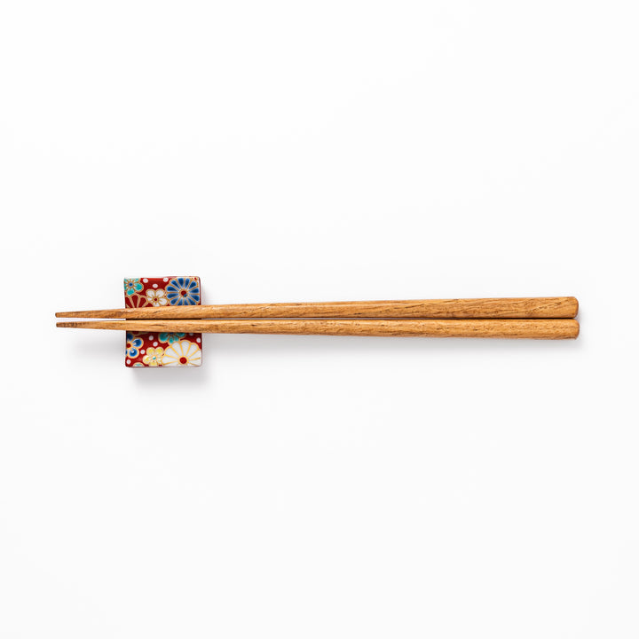 Rectangular Japanese ceramic chopstick rest with a red background decorated with colorful plum and chrysanthemum blossoms, perfect for elegant dining settings.