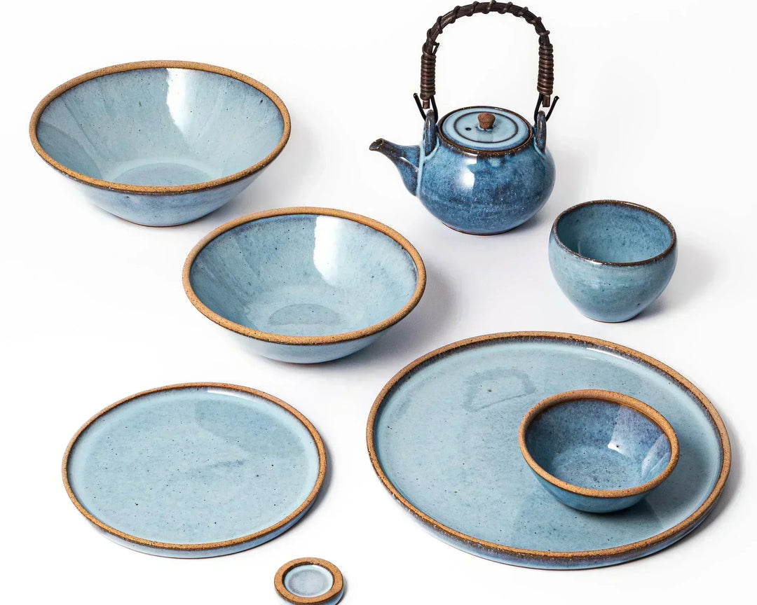 Blue handmade Japanese ceramic dish and teapot set with rustic brown rim showcasing bowls plates and cups on a white background