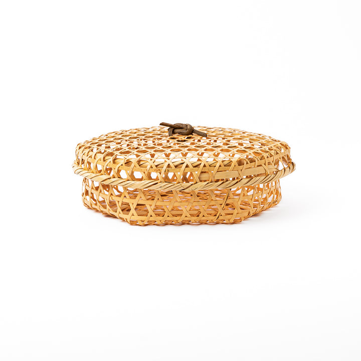 A closed round handwoven bamboo serving basket featuring a decorative brown knotted cord handle on top of the lid