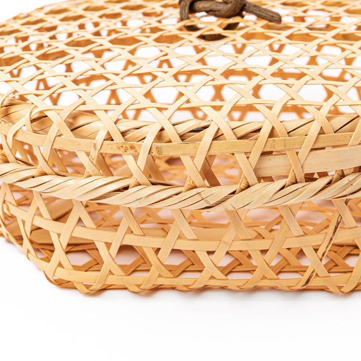 A closed round handwoven bamboo serving basket featuring a decorative brown knotted cord handle on top of the lid