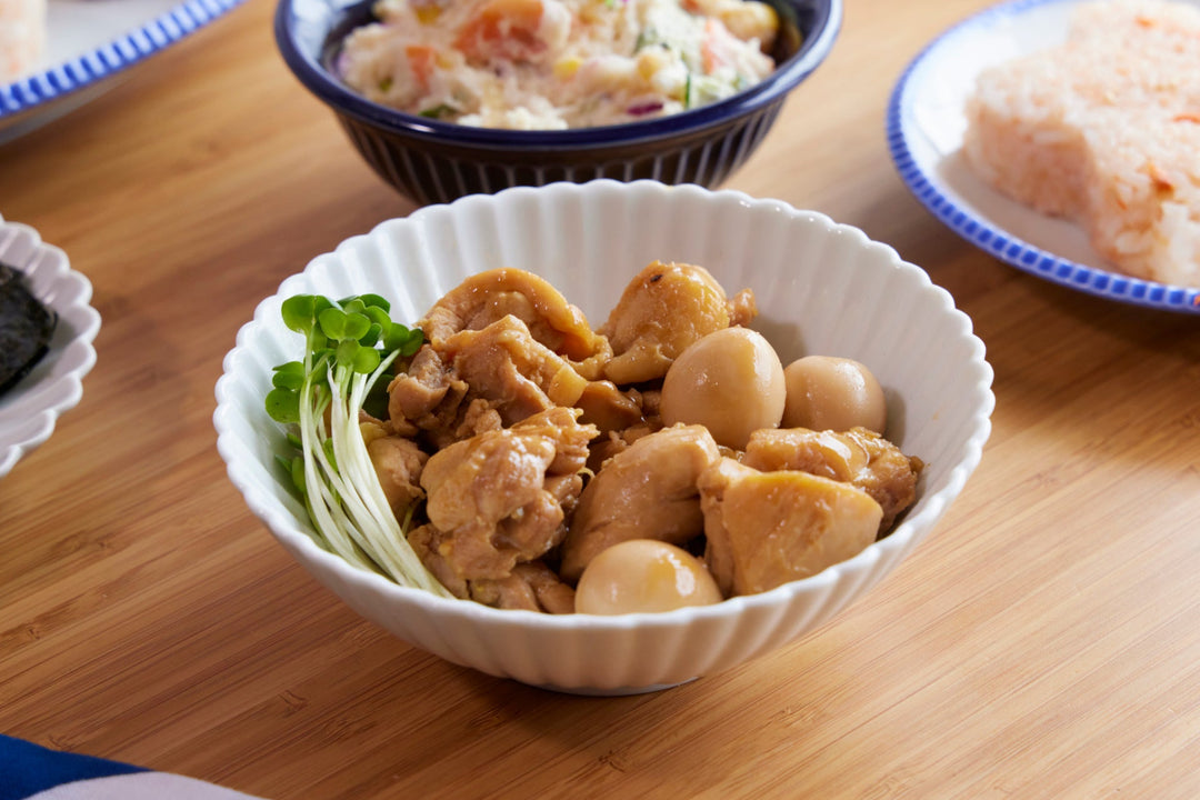 A white scalloped bowl filled with simmered chicken and quail eggs, served with microgreens on a wooden table during a home meal.