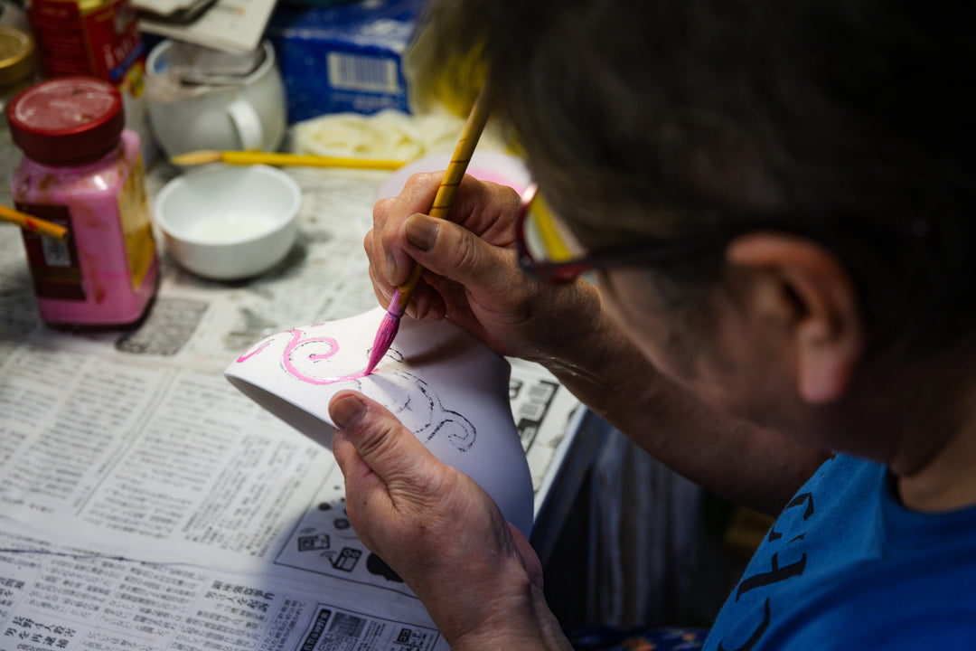 Artisan painting delicate pink details onto a ceramic dish at her workspace, showcasing traditional Japanese craftsmanship.