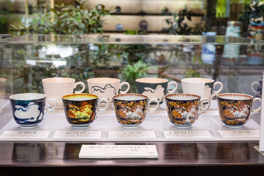 Showcase displaying porcelain cups from plain forms to intricately painted Imari-style pieces, arranged in a museum-like exhibit.